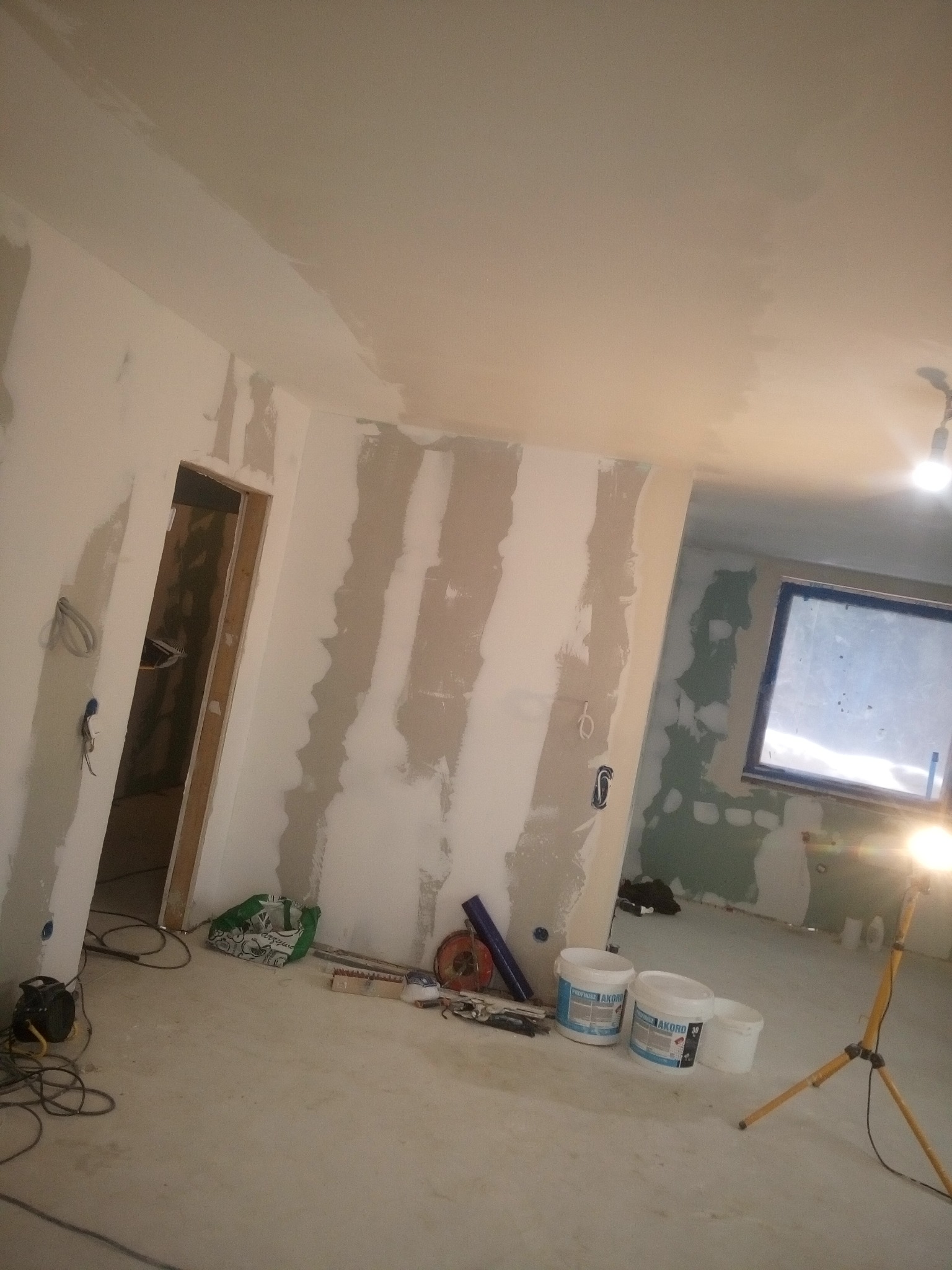 Wnętrze w trakcie prac przygotowawczych do wykończenia: ściany z fragmentami nałożonej gładzi, widoczne wiaderka z masą szpachlową Akord, przewody elektryczne wystające ze ścian, lampa budowlana...