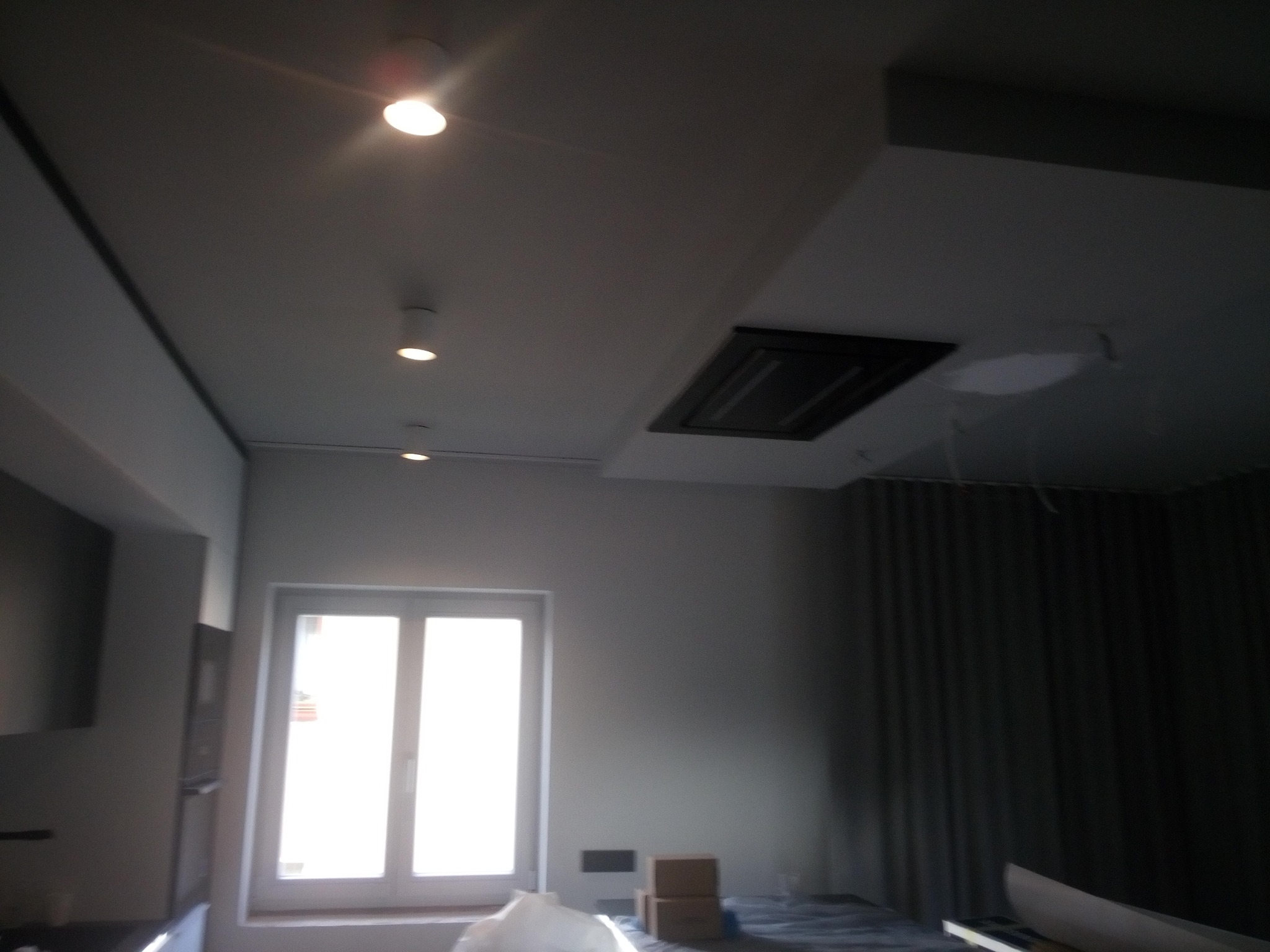 Wnętrze w trakcie wykańczania: widok na sufit z zamontowanym oświetleniem punktowym i wnęką, okno z widocznym światłem dziennym, zasłonięte okno oraz przygotowane materiały budowlane na podłodze.