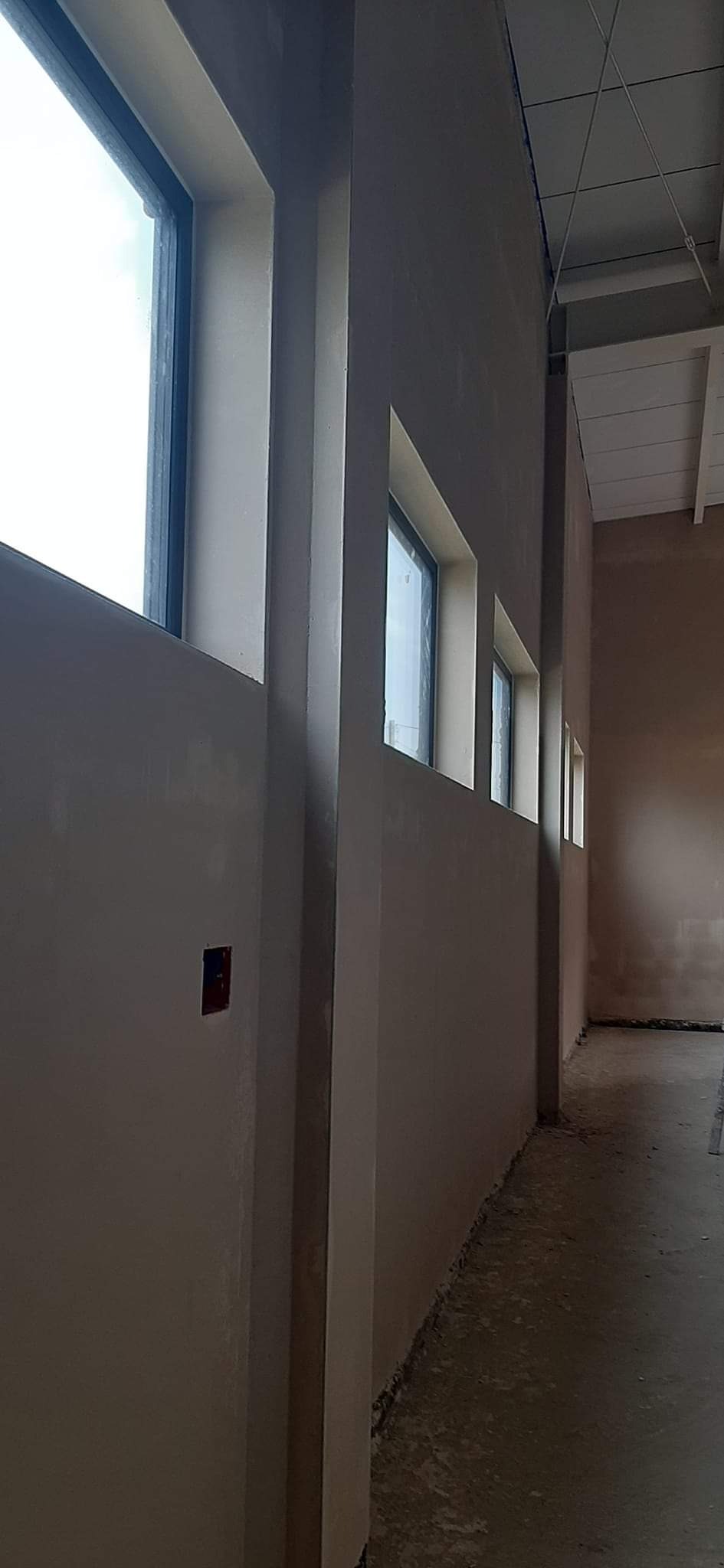 Świeżo otynkowana ściana wewnętrzna z rzędem okien, widoczne ślady prac, surowy beton na podłodze i fragment sufitu z instalacjami.