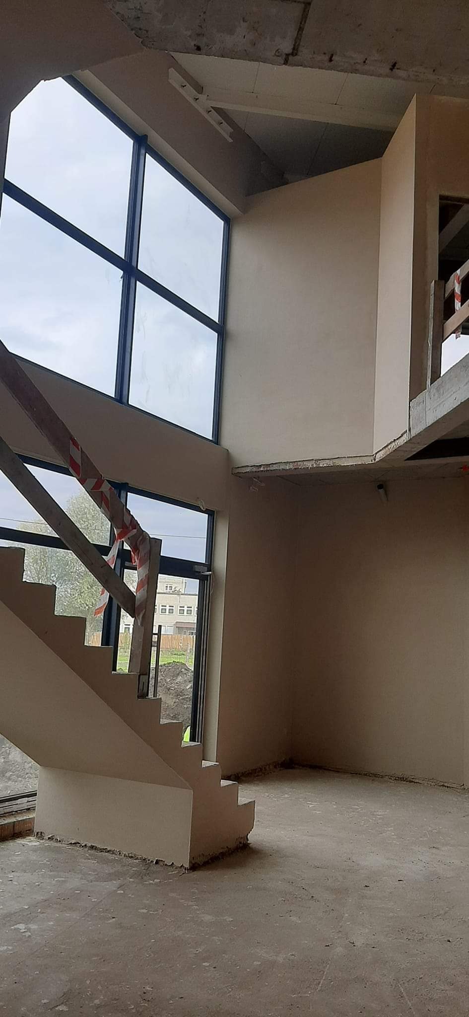 Wnętrze w trakcie prac wykończeniowych: świeżo otynkowane ściany, betonowa podłoga, schody z zabezpieczeniem taśmą ostrzegawczą, duże okna z widokiem na budynki.