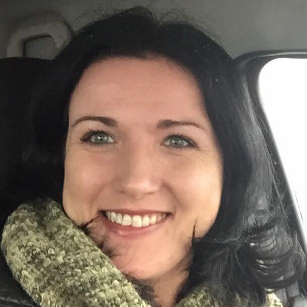 Portret uśmiechniętej kobiety o ciemnych włosach i zielonych oczach, w szaliku o grubym splocie, na neutralnym tle, z delikatnym oświetleniem.