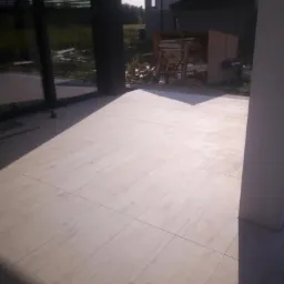 Jasne, kwadratowe płyty tarasowe ułożone na zewnątrz budynku, widoczne narzędzia i materiały budowlane w tle.