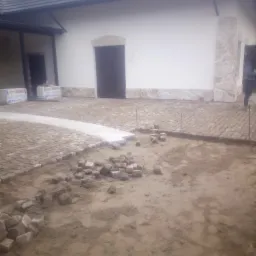 Fragment podwórka z częściowo ułożoną kostką brukową przed budynkiem z białymi ścianami i ciemnymi oknami, widoczne paczki styropianu oraz przygotowane kostki do dalszego układania.