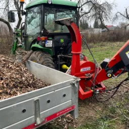 Ciągnik John Deere z przyczepą wypełnioną zrębkami drewna, obok czerwona rębarka do gałęzi, w tle drzewa bez liści i zabudowania.