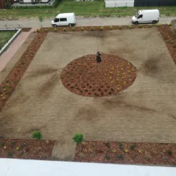 Widok z góry na świeżo założony trawnik z centralnym okręgiem z kory i nasadzeniami, obrzeża wyłożone korą i roślinami, dwa białe busy zaparkowane w tle.