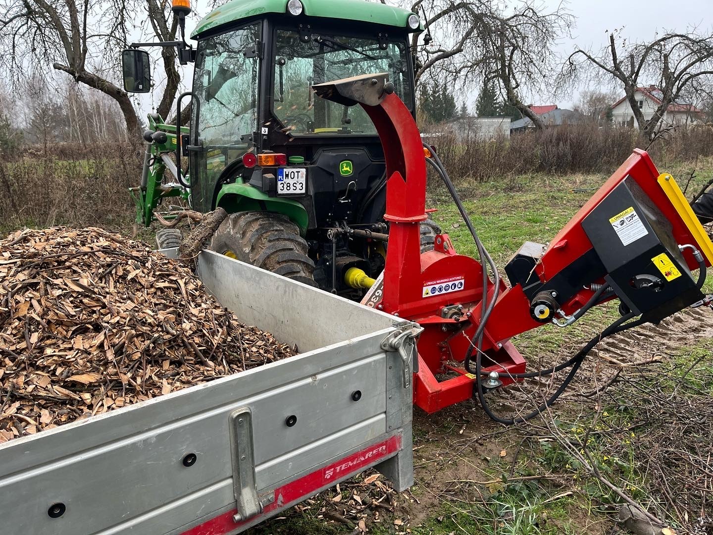 Ciągnik John Deere z przyczepą wypełnioną zrębkami drewna, obok czerwona rębarka do gałęzi, w tle drzewa bez liści i zabudowania.