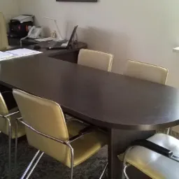 Ciemnobrązowy, duży stół konferencyjny z zaokrąglonymi bokami, otoczony jasnymi krzesłami na chromowanych nogach, w tle biurko z akcesoriami biurowymi i roślina doniczkowa na parapecie.