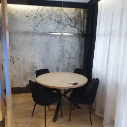 Jadalnia z okrągłym stołem z blatem imitującym jasne drewno, otoczonym czterema czarnymi krzesłami, na ścianie fototapeta z motywem drzewa bez liści, nad stołem nowoczesna lampa wisząca, obok okno...