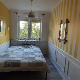 Sypialnia z żółtymi ścianami, jedna ściana w pionowe pasy, metalowe łóżko z patchworkową narzutą, dekoracyjne listwy ścienne i lustro w ozdobnej ramie.