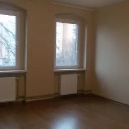 Pusty pok&oacute;j z dwoma oknami o zaokrąglonych g&oacute;rnych krawędziach, brązowymi panelami podłogowymi i beżowymi ścianami. Widoczne białe grzejniki pod oknami i fragment otwartego przejścia do innego...