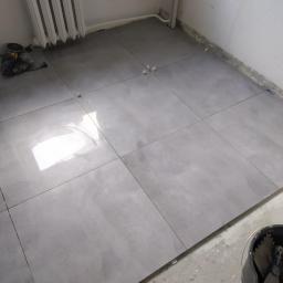 Michał Krypowicz - Szare płytki podłogowe ułożone w kwadraty, widoczne krzyżyki dystansowe, grzejnik żeberkowy przy ścianie, wiadro z narzędziami do układania płytek w rogu pomieszczenia.