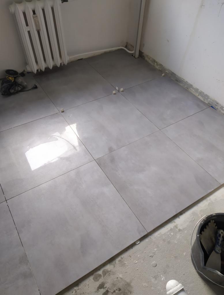 Szare płytki podłogowe ułożone w kwadraty, widoczne krzyżyki dystansowe, grzejnik żeberkowy przy ścianie, wiadro z narzędziami do układania płytek w rogu pomieszczenia.