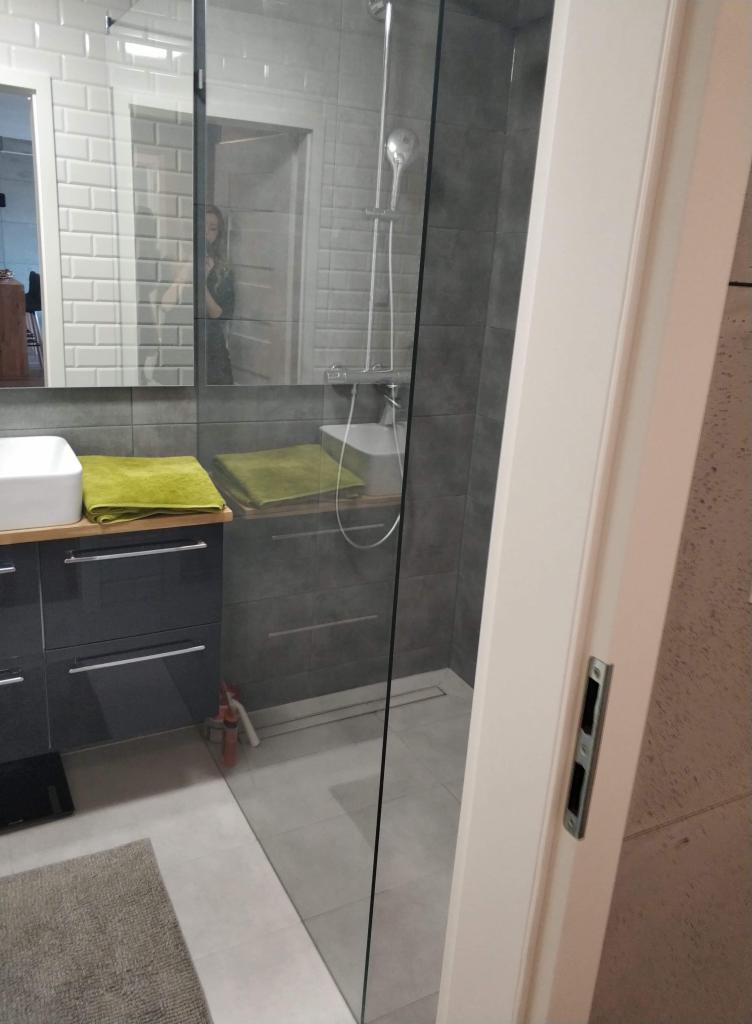 Nowoczesna łazienka z kabiną prysznicową typu walk-in, szarą glazurą, umywalką nablatową i zielonymi ręcznikami, widok zza uchylonych drzwi.