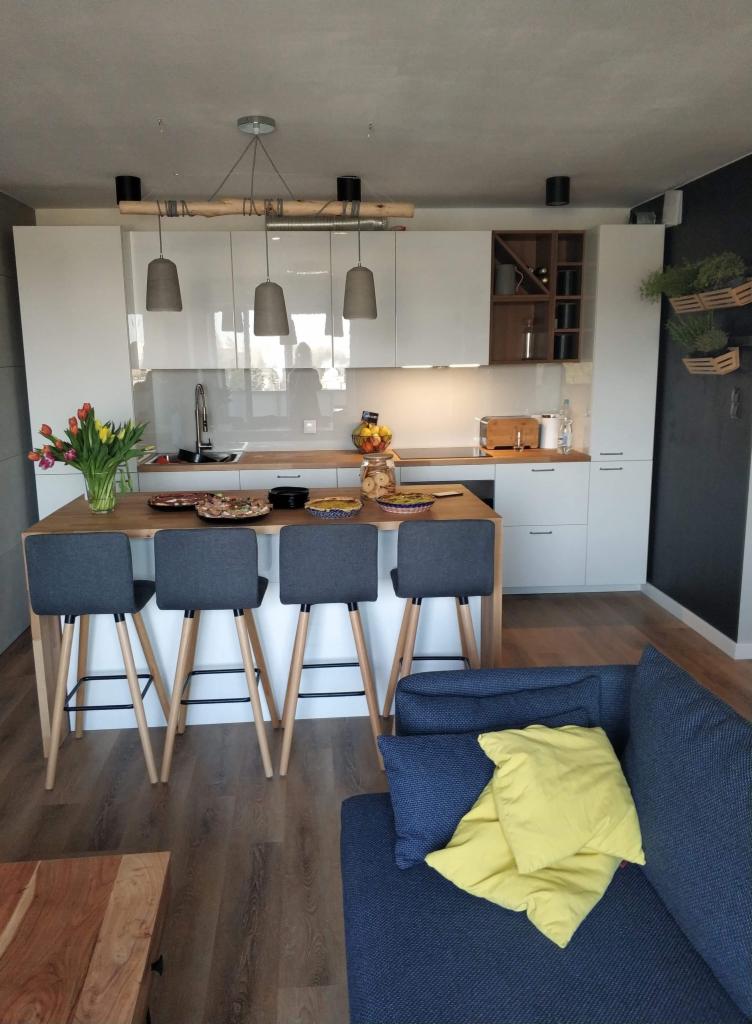 Nowoczesna kuchnia z wyspą i czterema szarymi hokerami, na blacie wyspy talerze z przekąskami, nad wyspą designerska lampa z drewnianym elementem, w tle białe szafki i fragment salonu z niebieską...