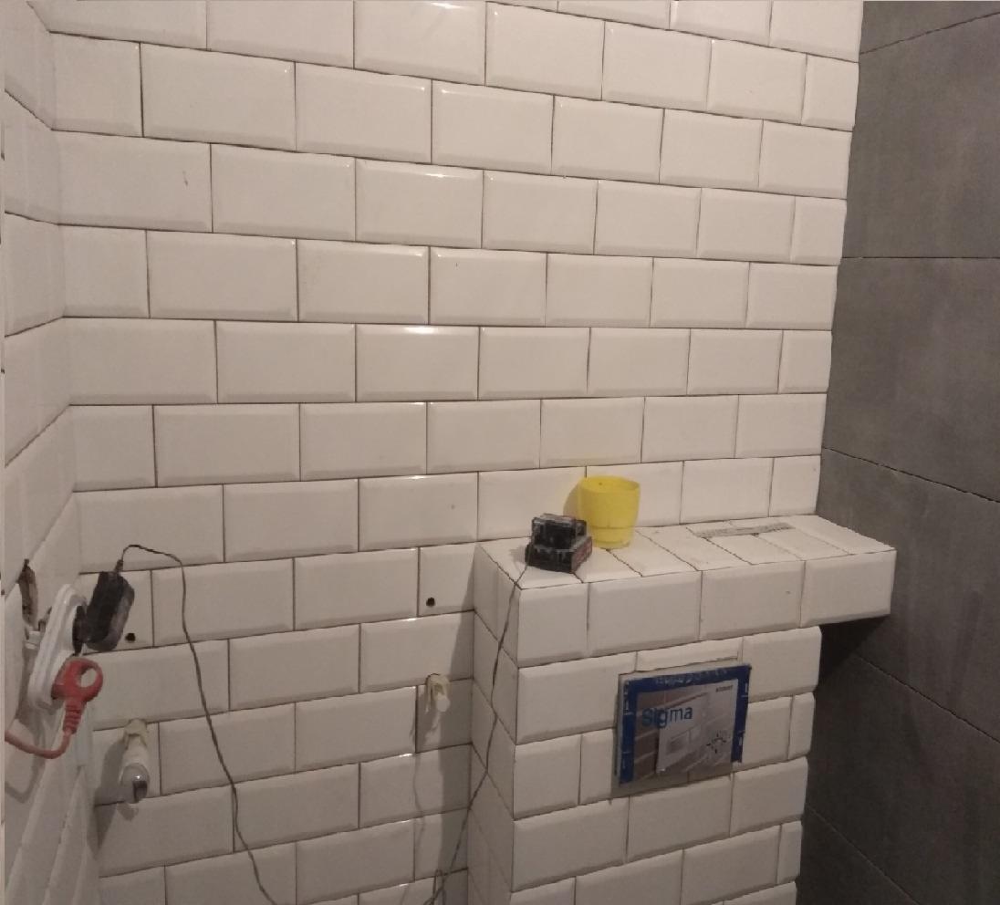 Ściana łazienki wyłożona białymi, prostokątnymi płytkami typu metro, z widocznymi przewodami elektrycznymi, puszką podtynkową Sigma i żółtym kubkiem na półce z płytek.