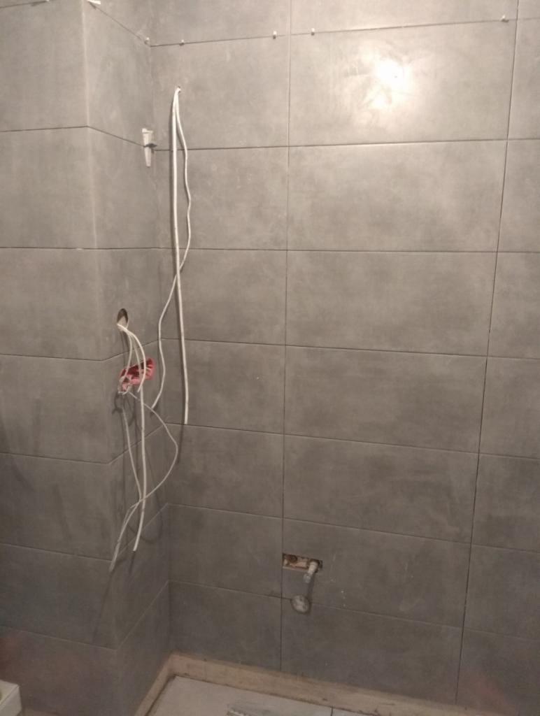 Ściana łazienki w trakcie układania szarych, prostokątnych płytek, widoczne wystające przewody elektryczne i rury hydrauliczne przed podłączeniem armatury.