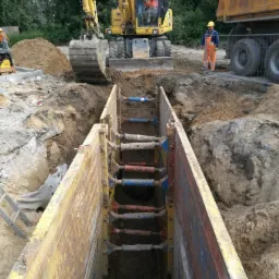 Żółty wykop z zabezpieczeniami, koparka i ciężarówka w tle, pracownicy w odblaskowych kombinezonach, przygotowanie do instalacji podziemnych.