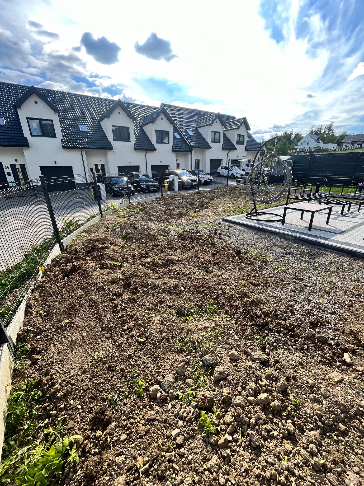 Przygotowany teren pod przyszły ogród przed szeregiem nowoczesnych domów, widoczne świeżo przekopana ziemia, fragment trawnika i meble ogrodowe.