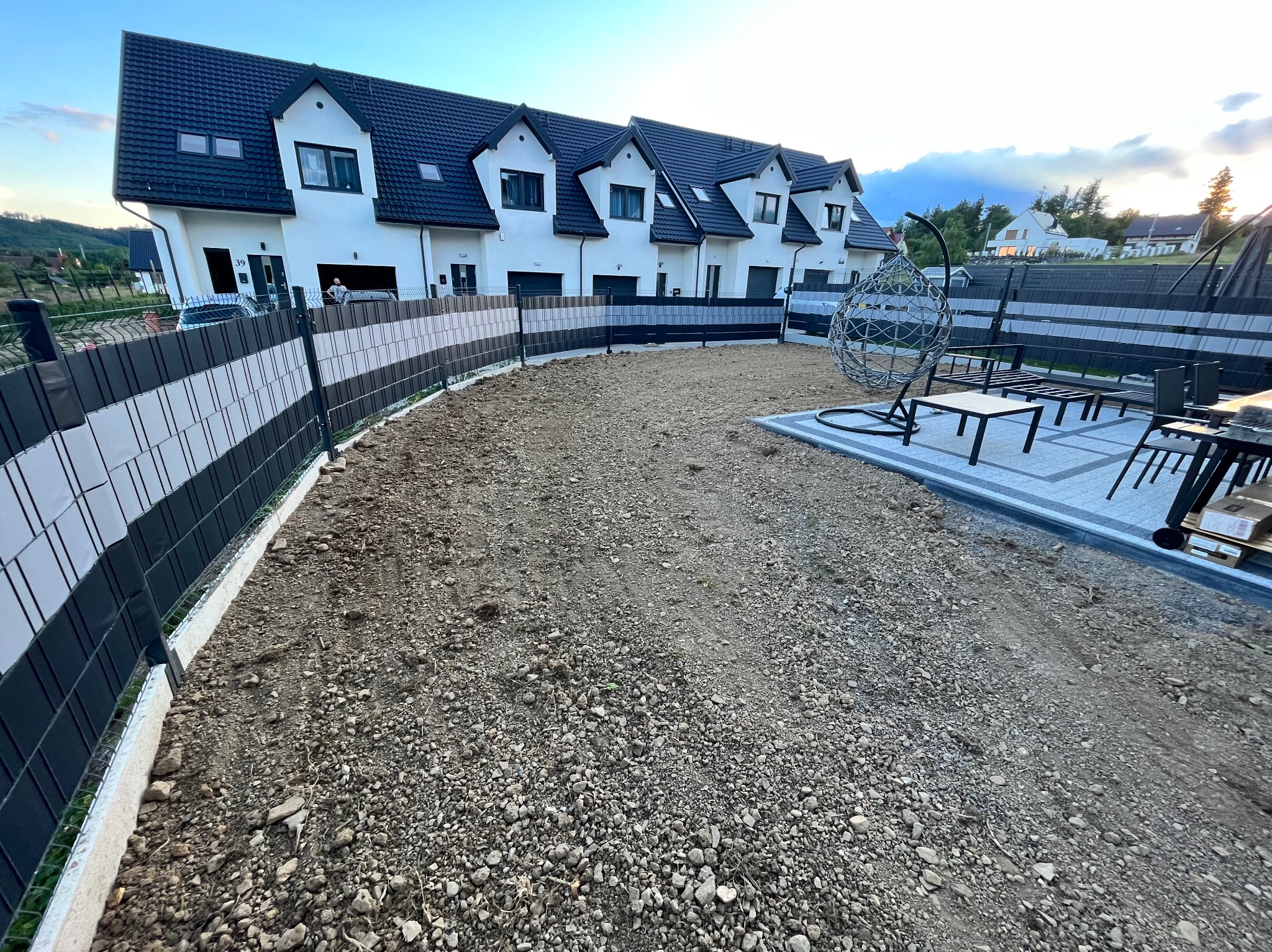 Przygotowany teren pod ogród, z widocznym ogrodzeniem panelowym, meblami ogrodowymi i nowymi segmentami domów w tle.