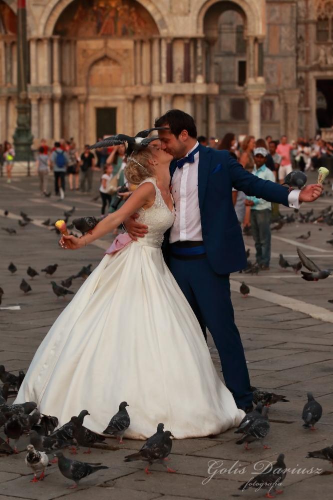 Pocałunek nowożeńców na tle weneckiej architektury, otoczonych gołębiami i trzymających lody.