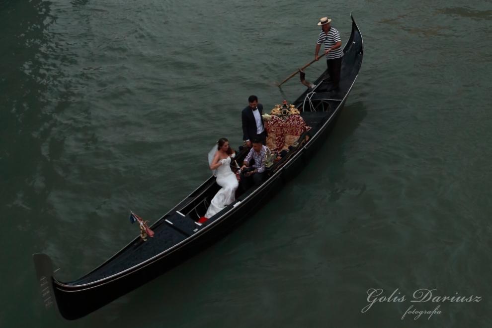 Gondola w Wenecji z parą młodą w strojach ślubnych, muzyk grający na instrumencie i gondolier w paski, widok z góry.