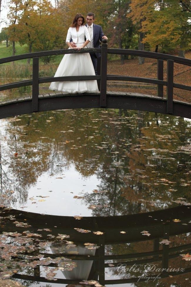 Para młoda w jesiennej scenerii stoi na drewnianym moście, którego odbicie w wodzie tworzy malowniczy efekt.