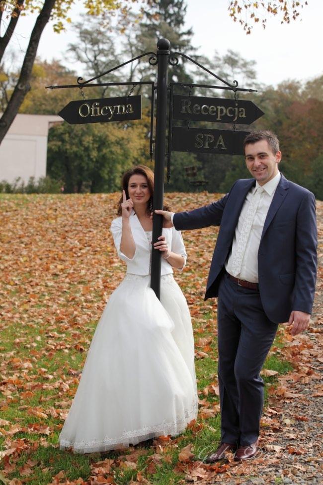 Para młoda w plenerze pozuje przy drogowskazie z napisem 'Oficyna' i 'Recepcja SPA', otoczona opadłymi liśćmi.