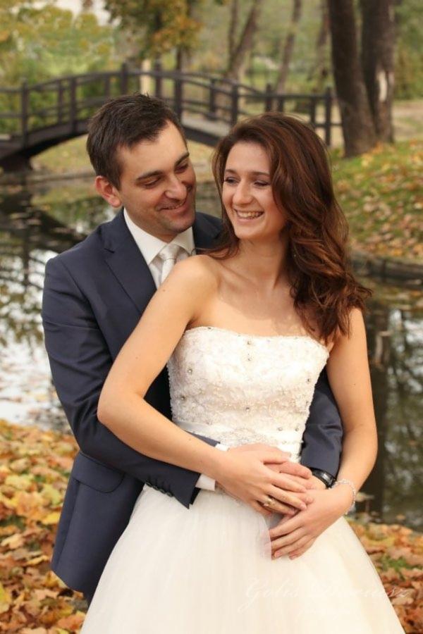 Uśmiechnięta para młoda w objęciach na tle parku z mostkiem i stawem, jesienne liście na ziemi.