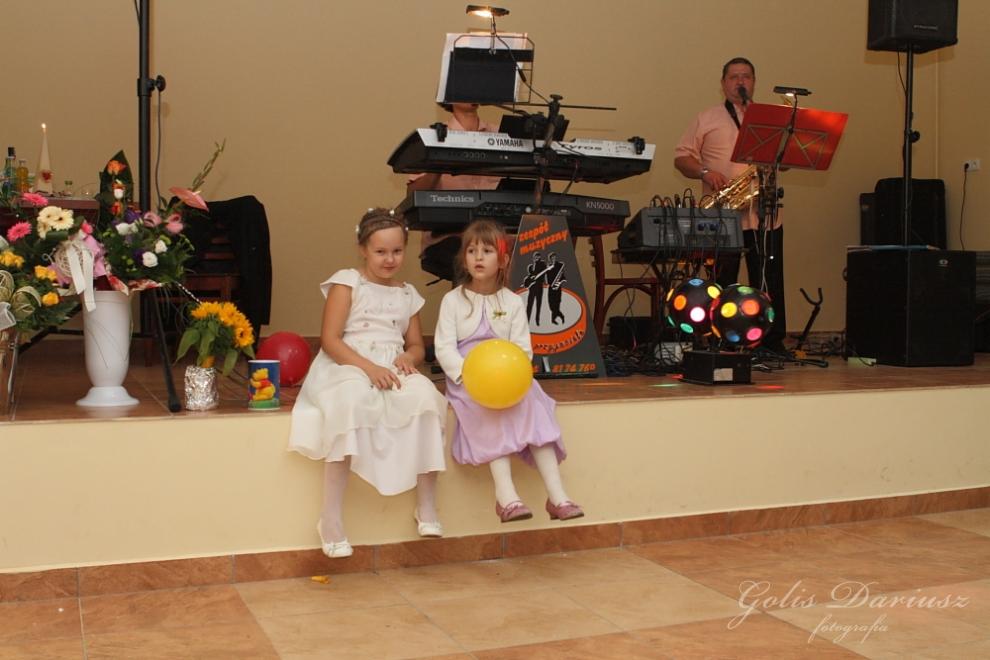 Dwie dziewczynki w sukienkach siedzą na krawędzi sceny podczas imprezy z zespołem muzycznym grającym na instrumentach klawiszowych i saksofonie, dekoracje kwiatowe i kolorowe kule na scenie.