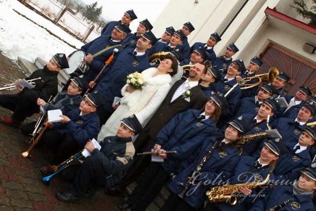 Panna młoda z bukietem i orkiestra dęta w granatowych mundurach pozują przed kościołem w pochmurny dzień.