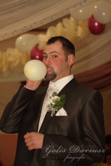 Pan młody w ciemnym garniturze z butonierką dmucha biały balon na tle dekoracji sali weselnej z balonami.