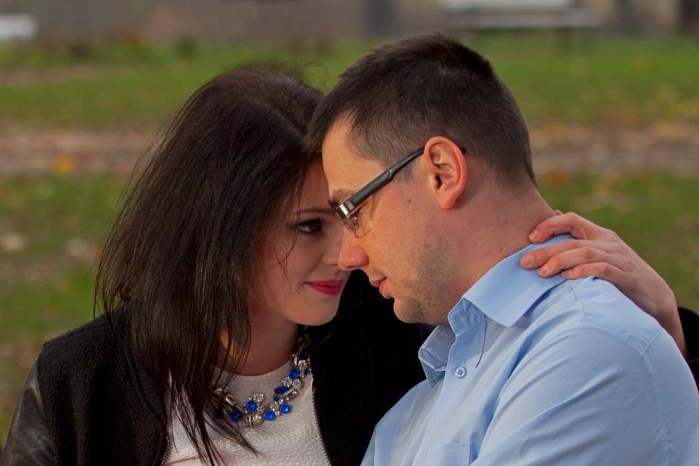 Zakochana para, kobieta w białej sukience i naszyjniku z niebieskimi kamieniami obejmuje mężczyznę w okularach, zbliżając się twarzami na tle zieleni.