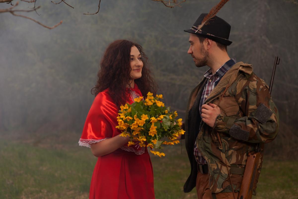 Para w leśnej scenerii: kobieta w czerwonej sukni z bukietem żółtych kwiatów, mężczyzna w kapeluszu z piórem i kurtce moro, trzymający strzelbę.