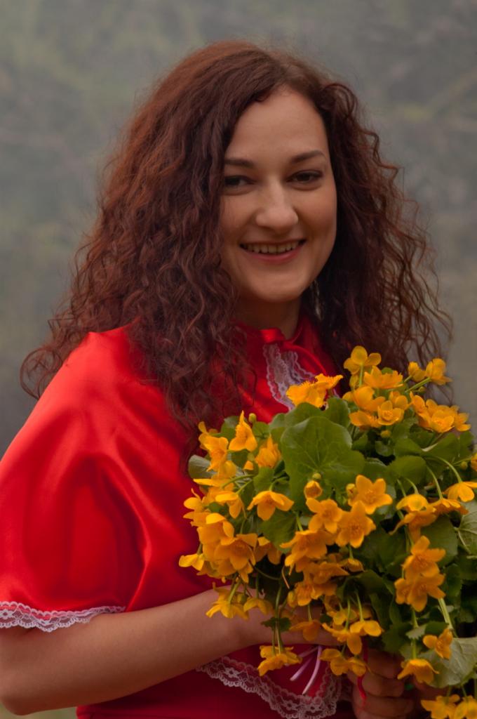 Uśmiechnięta kobieta z kręconymi kasztanowymi włosami, ubrana w czerwoną bluzkę z koronkowym wykończeniem, trzyma bukiet jaskrawożółtych kaczeńców na tle rozmytego krajobrazu.