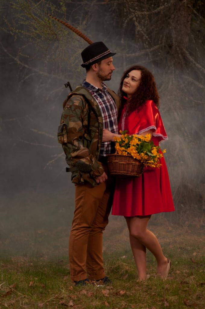 Stylizowana sesja zdjęciowa pary: mężczyzna w stroju myśliwego z piórem przy kapeluszu i kobieta przebrana za Czerwonego Kapturka z koszykiem żółtych kwiatów, na tle leśnej scenerii z mgłą.