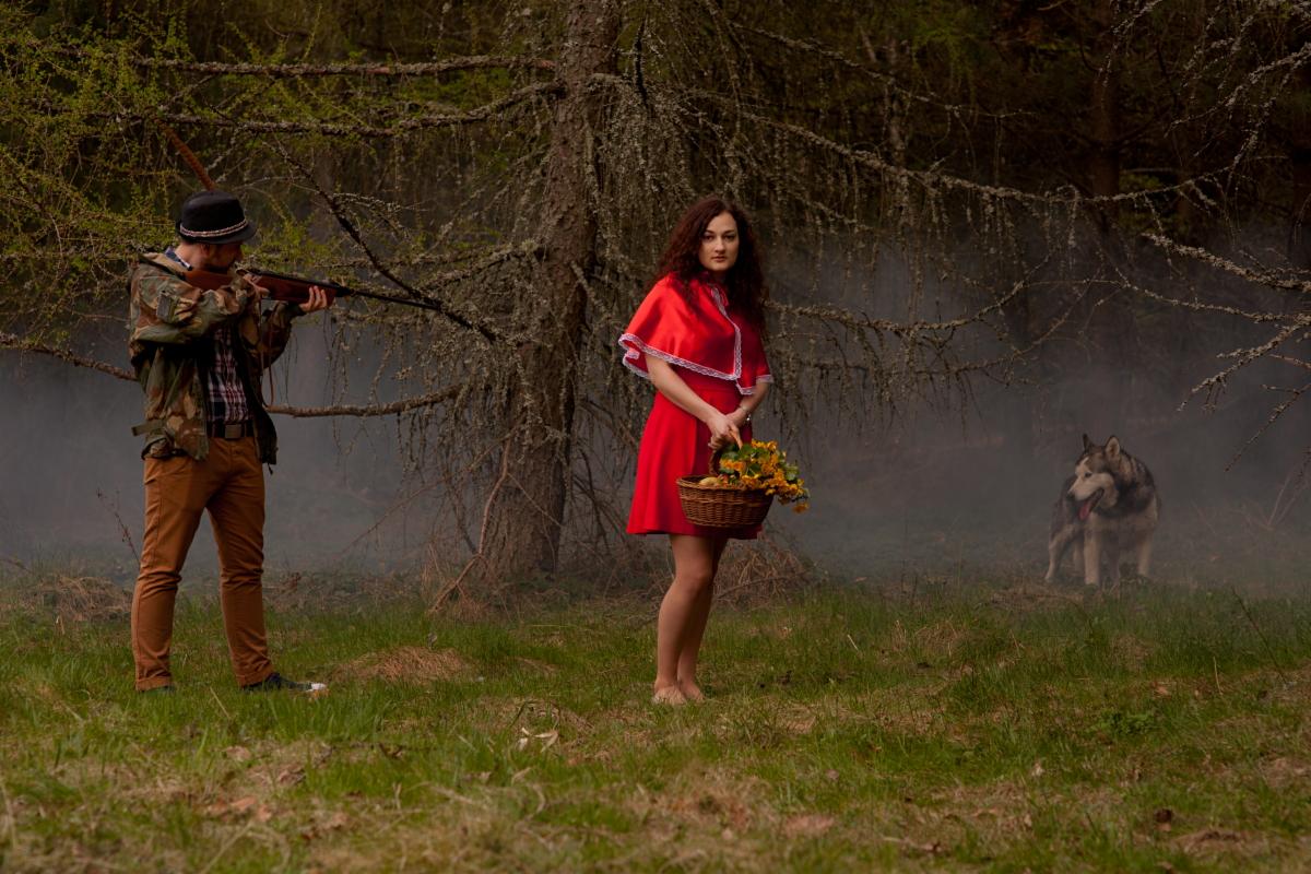 Stylizowana sesja zdjęciowa: Kobieta w czerwonej sukience z koszykiem kwiatów, mężczyzna celujący z broni, w tle wilczasty pies w zalesionym terenie z mgłą.