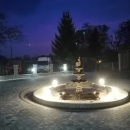 Oświetlona fontanna na posesji wyłożonej szarą kostką brukową, widok wieczorny, w tle zaparkowany biały bus.