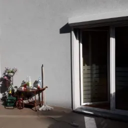 Dwa okna PCV z roletami zewnętrznymi, jedno otwarte, drugie zamknięte, na balkonie z kwiatami i stolikiem na kółkach, widoczne cienie