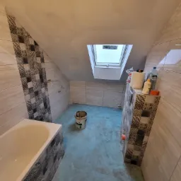 Łazienka na poddaszu w trakcie prac wykończeniowych: położone płytki na ścianach, niebieska hydroizolacja na podłodze, widoczne narzędzia i materiały budowlane, okno dachowe.