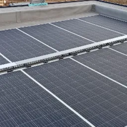 Widok z góry na panele fotowoltaiczne zamontowane na płaskim dachu z szarą, metalową konstrukcją mocującą pośrodku, z widocznym fragmentem elewacji budynku po prawej stronie.