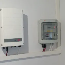 Biały inwerter solarny marki SolarEdge zamontowany obok skrzynki z bezpiecznikami na białej ścianie, z widocznymi kablami i listwą maskującą.