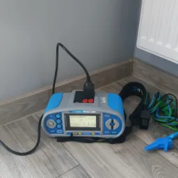 Miernik elektryczny podłączony do gniazdka, z widocznymi przewodami pomiarowymi i zaciskami krokodylkowymi na podłodze z paneli.