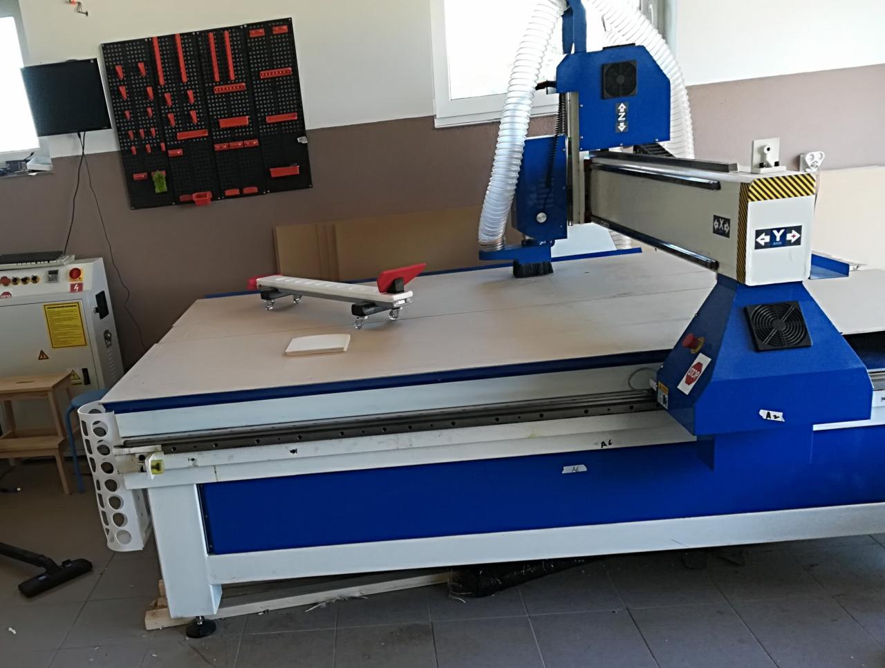 Duża frezarka CNC z niebieską obudową, widoczny panel sterowania, listwa z narzędziami na ścianie w tle.