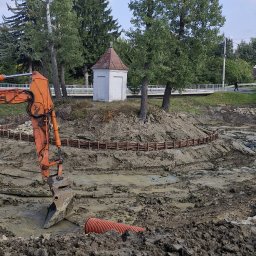 Jarosław Florek Koparki - Pomarańczowa koparka w błotnistym wykopie, widoczny fragment rury. W tle mała kapliczka na wzgórzu otoczona drzewami i drewnianym płotkiem.