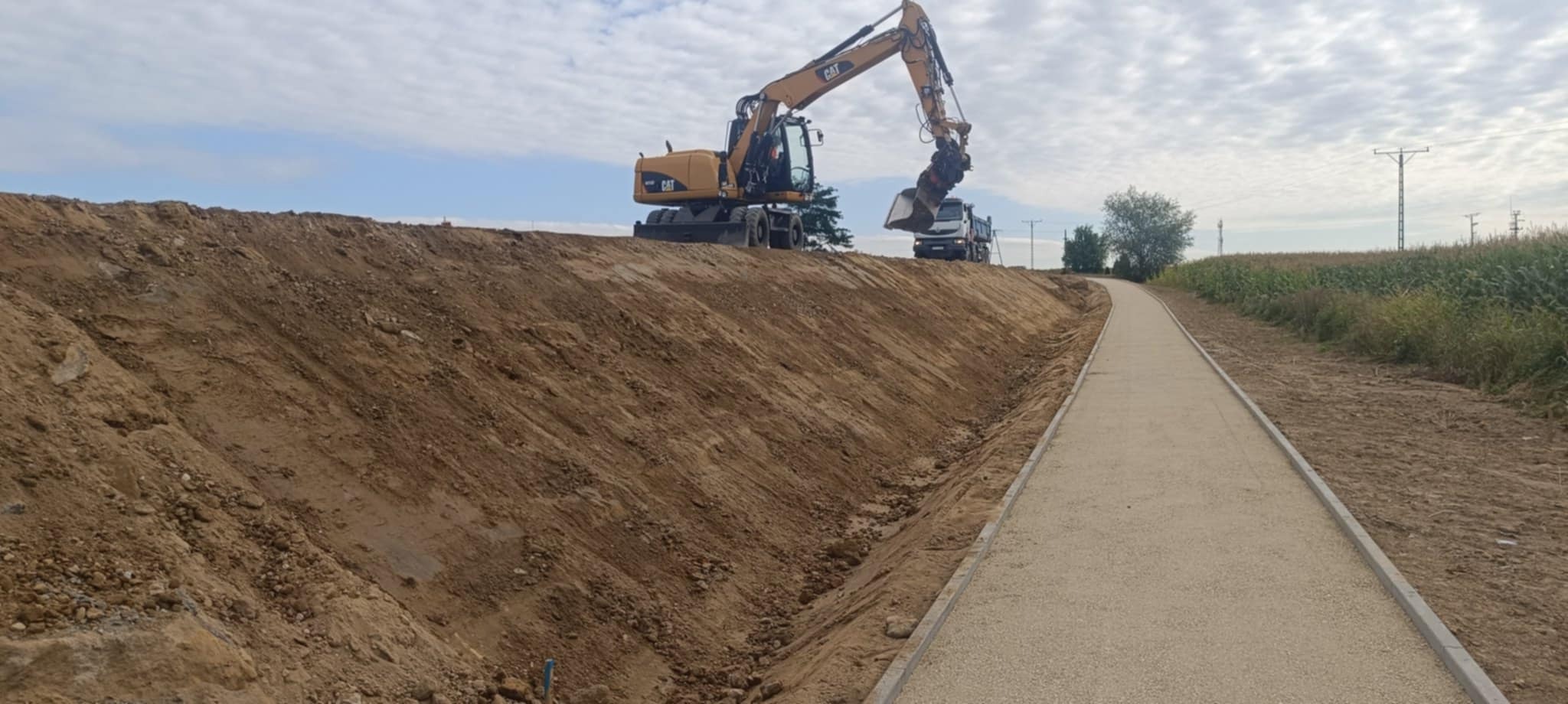 Żółta koparka CAT nabiera ziemię z wykopu, obok utwardzona droga z kostką. W tle ciężarówka i pole kukurydzy pod pochmurnym niebem. Prace ziemne.