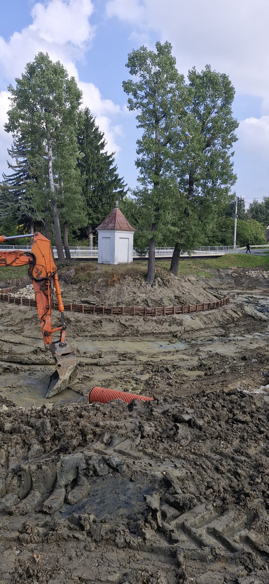 Pomarańczowa koparka w błotnistym wykopie, widoczny fragment rury. W tle mała kapliczka na wzgórzu otoczona drzewami i drewnianym płotkiem.