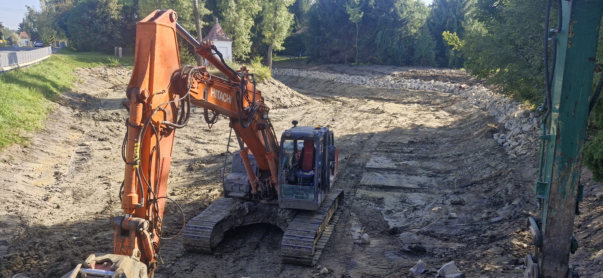 Pomarańczowa koparka Hitachi w trakcie prac ziemnych przy korycie rzeki, z widocznymi brzegami umocnionymi kamieniami. Maszyna na tle zieleni drzew.