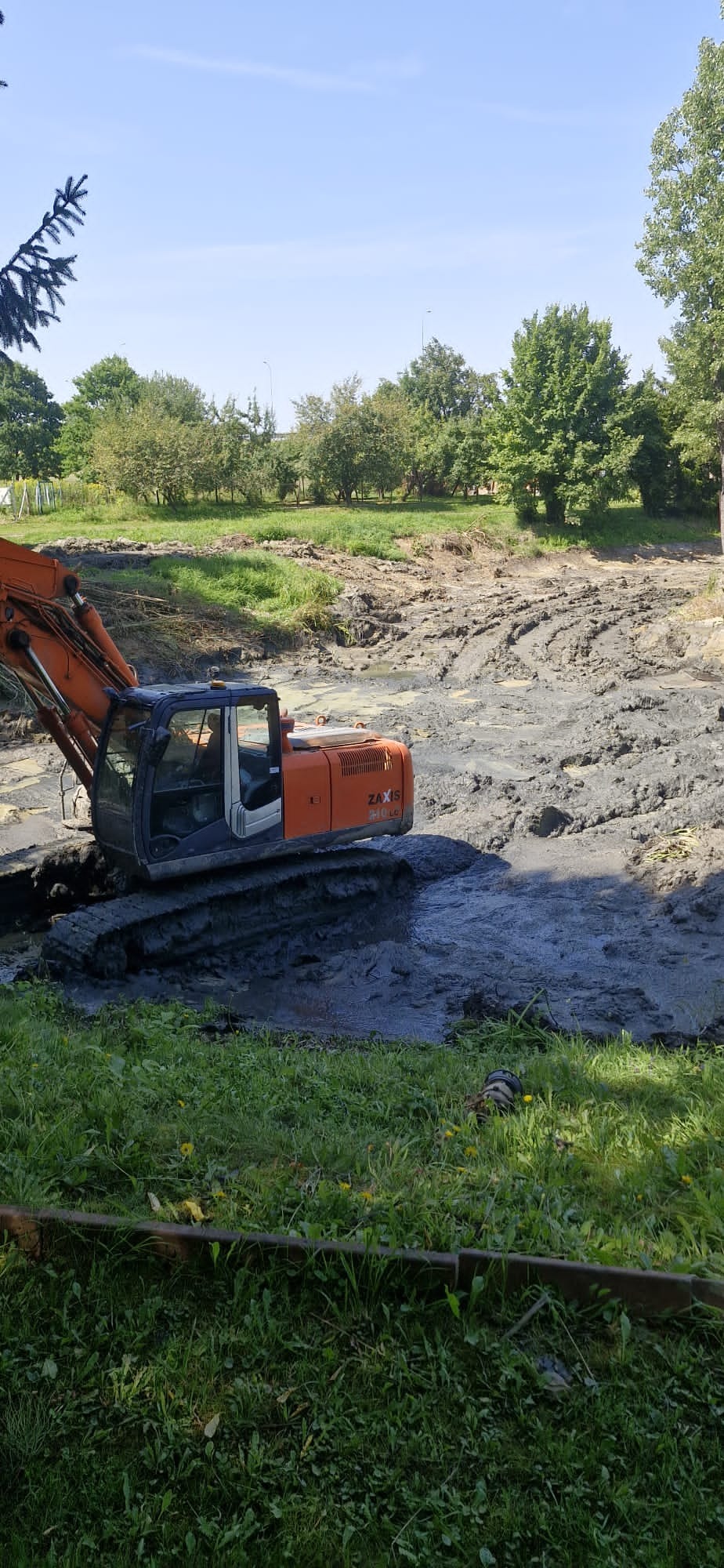 Pomarańczowa koparka Zaxis w błotnistym wykopie, otoczona zielenią drzew i trawy, w słoneczny dzień. Widoczne ślady opon w błocie.