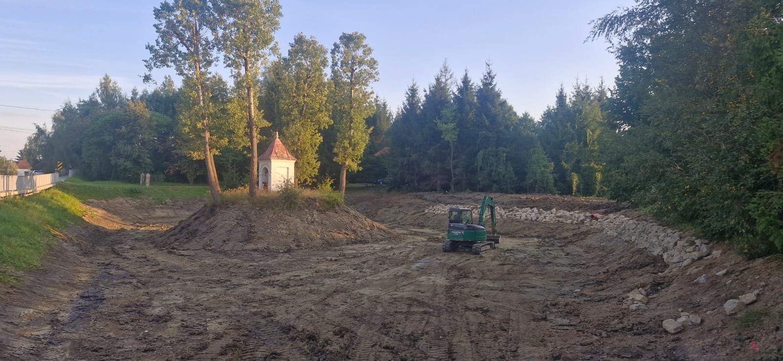 Wykopany staw z wysepką i kapliczką w tle, zabezpieczone brzegami z kamieni. Na pierwszym planie mała koparka. Widok na zalesiony teren.