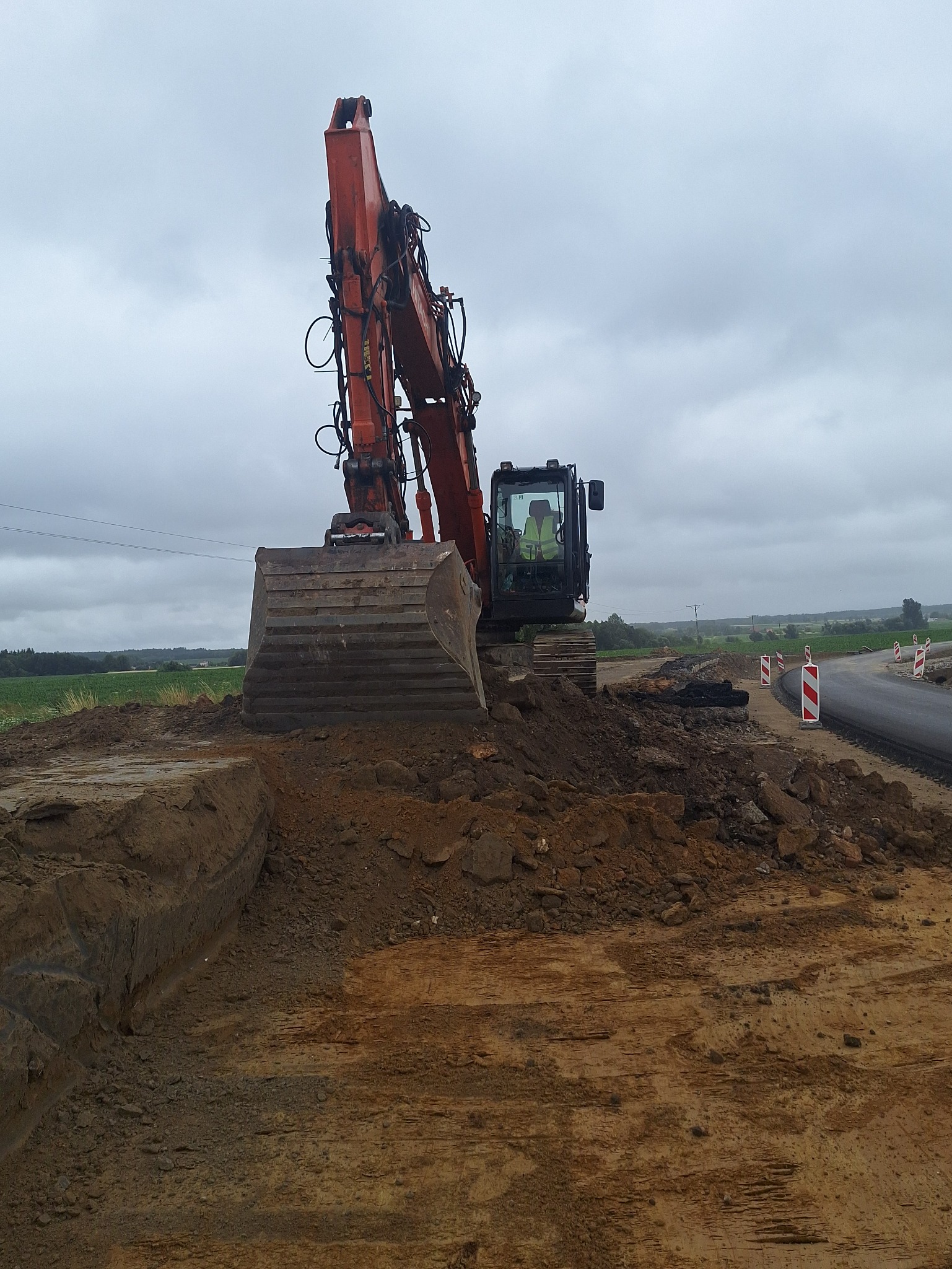 Pomarańczowa koparka na placu budowy drogi, widoczne bariery drogowe i operator w kabinie, pochmurne niebo w tle. Wykop ziemi.